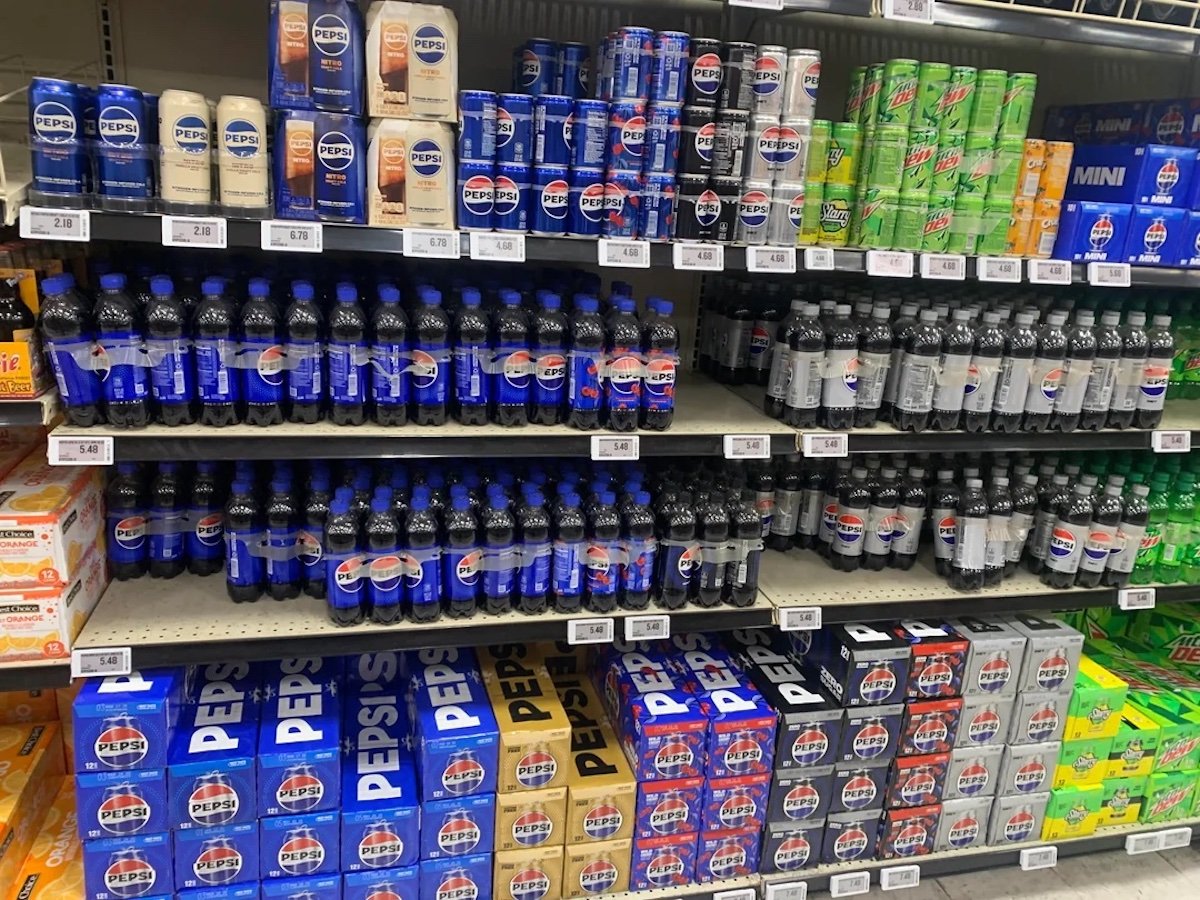 Shelves in a grocery store stocked with various soft drink brands, including Pepsi, Diet Pepsi, Mountain Dew, and Sierra Mist in bottles and cans, with price tags visible below each row.