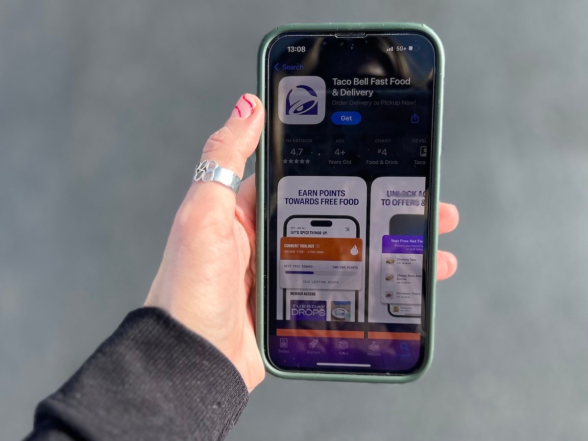 A hand with a silver ring holds a smartphone displaying the Taco Bell Fast Food & Delivery app page in the App Store. The screen shows app details and preview images.