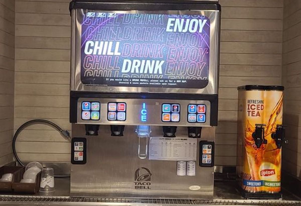 A Taco Bell self-serve drink station with soda dispensers, ice dispenser, cup lids, straws on the left, and a Lipton iced tea dispenser on the right. The sign above reads "CHILL DRINK ENJOY" in colorful letters.