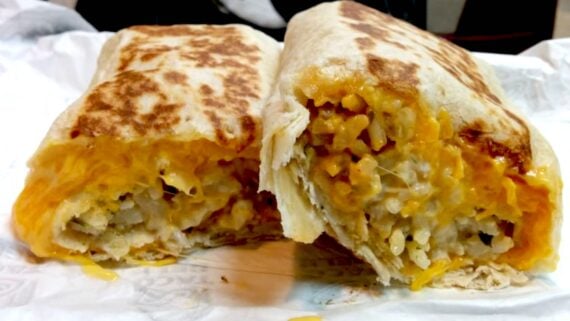 A close-up of a burrito cut in half, showing a filling of melted cheese and diced potatoes wrapped in a grilled flour tortilla, resting on white paper.