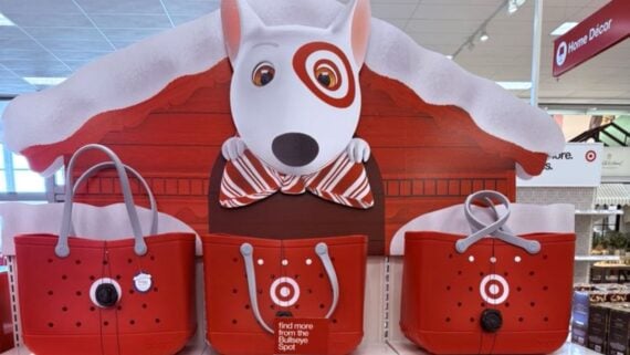 A store display features Target's bull terrier mascot peeking over a red doghouse with snow on the roof. Below are three red tote bags with the Target bullseye logo, set on white shelves in a store.