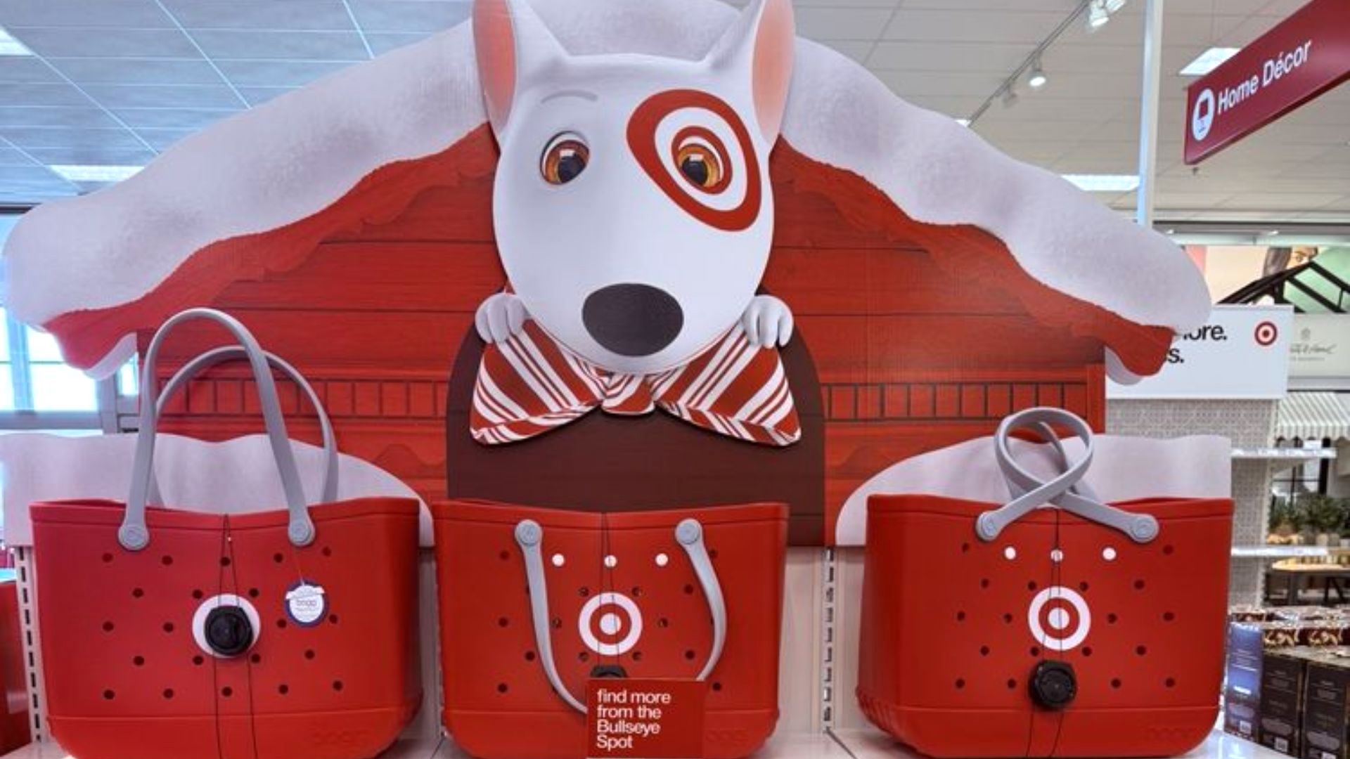 A store display features Target's bull terrier mascot peeking over a red doghouse with snow on the roof. Below are three red tote bags with the Target bullseye logo, set on white shelves in a store.