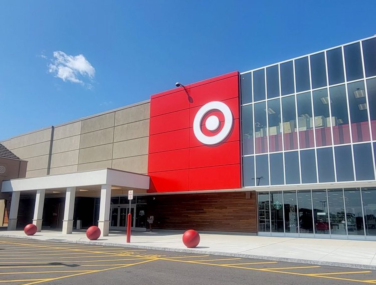 A Target store exterior on a sunny day, featuring the red Target logo on a large red panel, glass windows, entrance doors, and red bollards in front of the building.