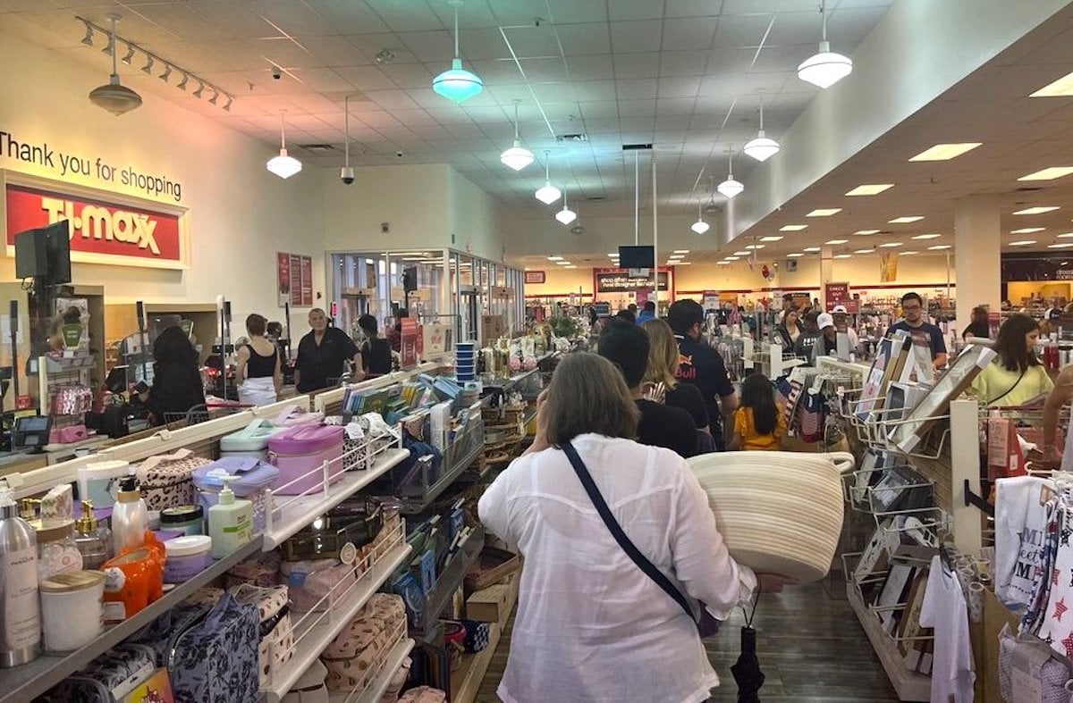 Shoppers stand in a long checkout line inside a busy TJ Maxx store filled with home goods, decor, and accessories. Bright lighting and crowded aisles create a bustling atmosphere.