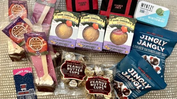 An assortment of Trader Joe's holiday treats, including chocolate stirring spoons, dark chocolate orange, peanut butter cups, Jingly Jangly snack mix, wintermint gum, and a festive matchbox, displayed on a textured surface.
