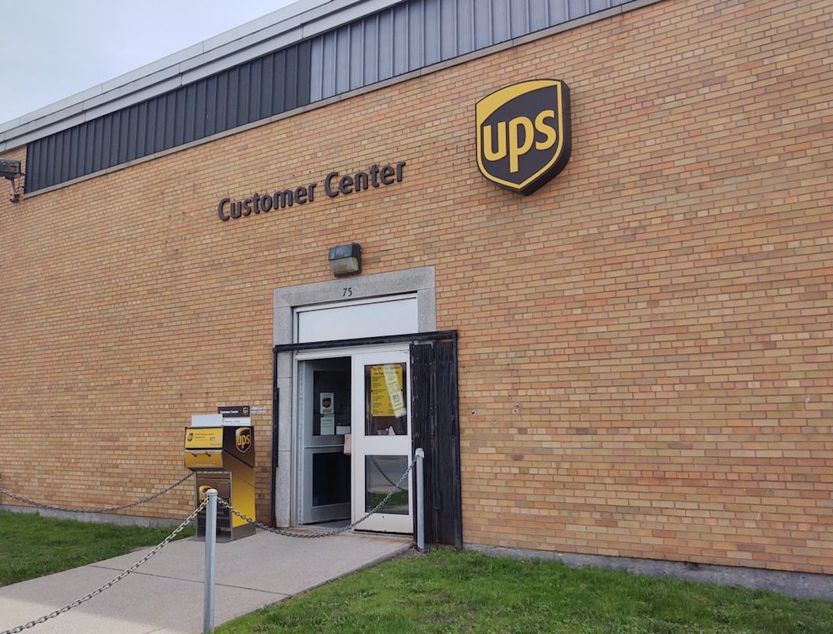 A UPS Customer Center with a brown brick exterior, a UPS sign above the entrance, and a yellow UPS drop box near the doorway. Grass and a sidewalk are in the foreground.