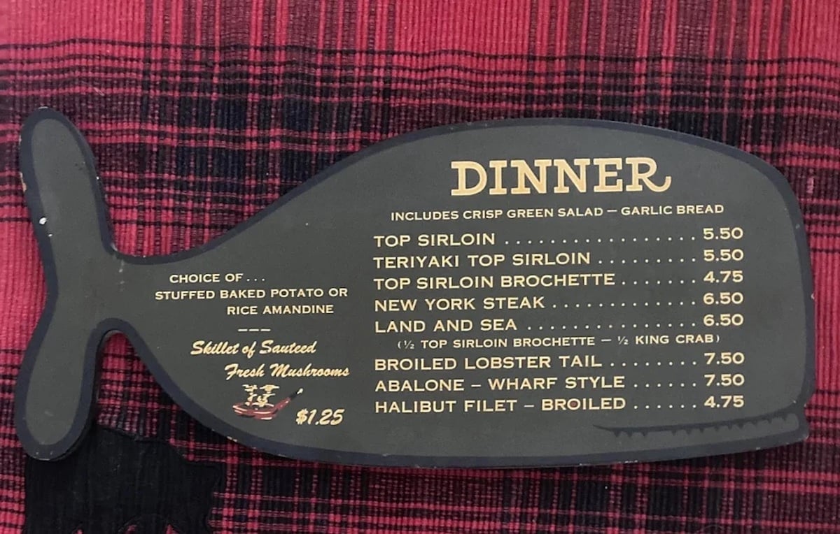 A fish-shaped black menu lists dinner options and prices, including various sirloins, NY steak, lobster tail, abalone, and halibut. Dinners include salad and garlic bread; sides and mushrooms are also available. Menu lies on red patterned cloth.