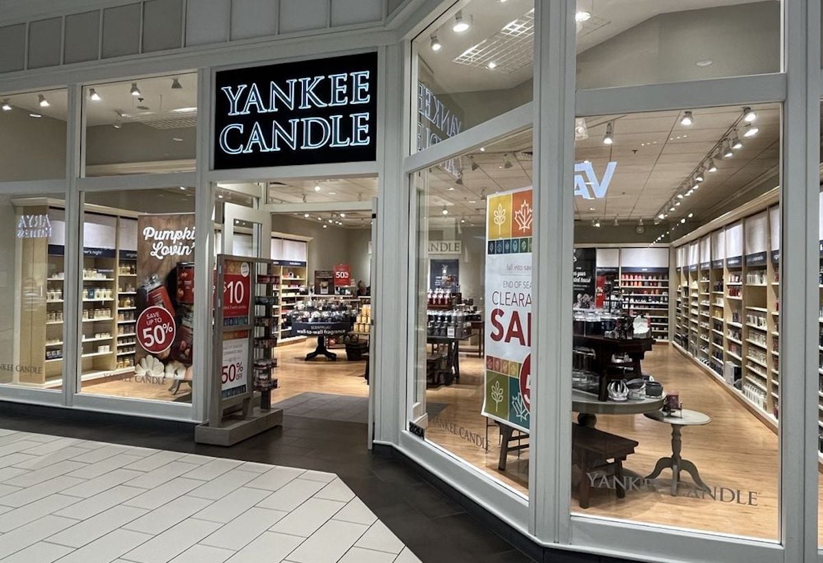 A Yankee Candle store in a mall with large glass windows. Signs advertise a clearance sale, 50% off, and seasonal promotions. Shelves inside are stocked with various candles and products. The store looks bright and inviting.