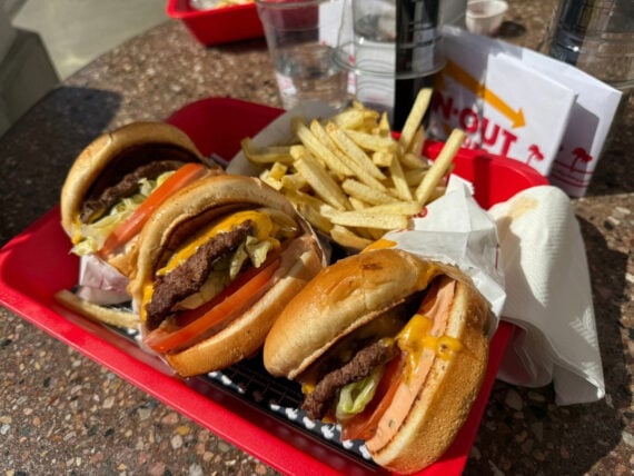 A red tray holds two cheeseburgers with lettuce, tomato, and cheese, alongside a serving of French fries. Napkins, a drink, and an In-N-Out menu are in the background on a speckled table.