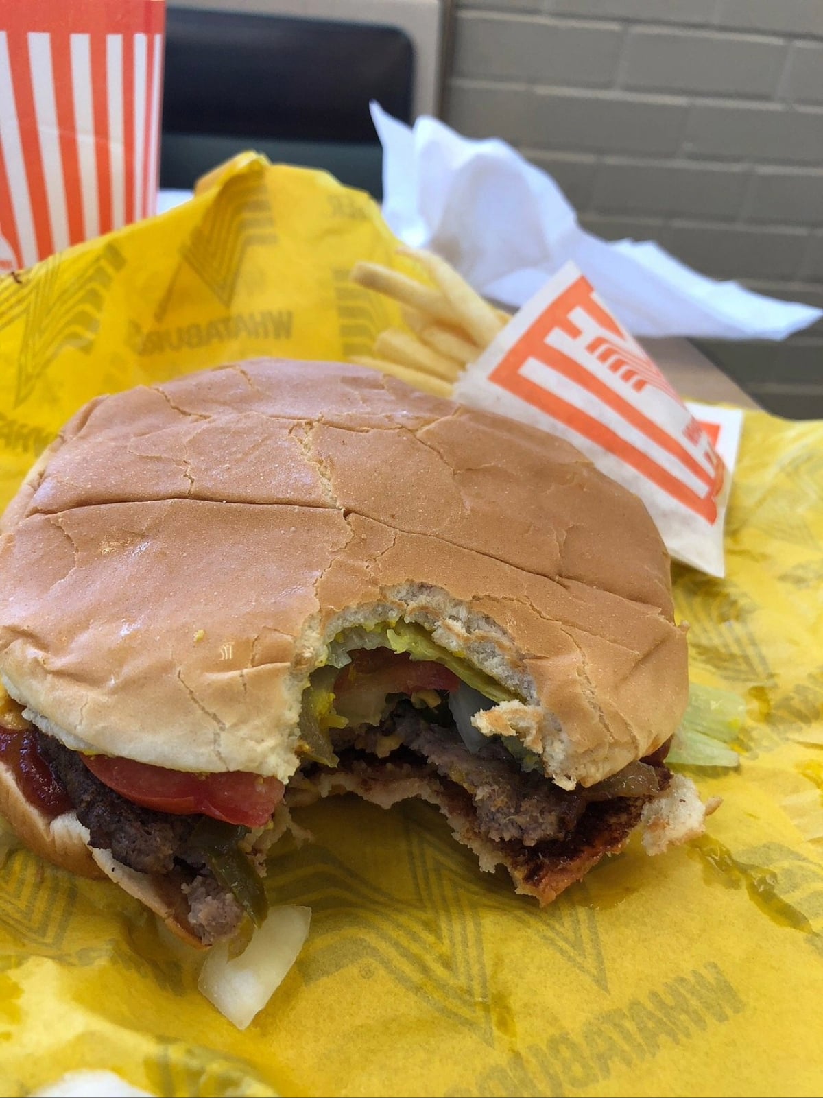 A Whataburger hamburger with a bite taken out, showing lettuce, tomato, jalapeños, and beef patty, sits on yellow Whataburger paper with a serving of fries and a drink in the background.