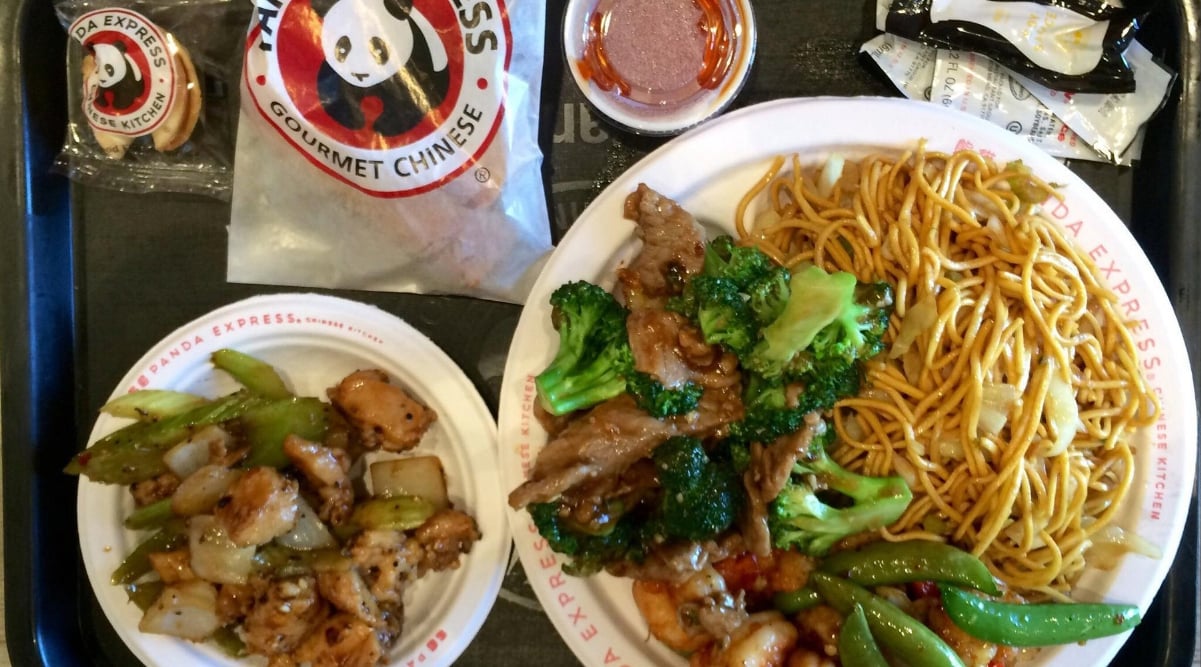 A tray with Panda Express food—a favorite among West Coast fast-food chains—featuring a large plate of noodles, beef with broccoli, mixed vegetables, a smaller plate of chicken and veggies, fortune cookies, soy sauce, and a container of sauce.