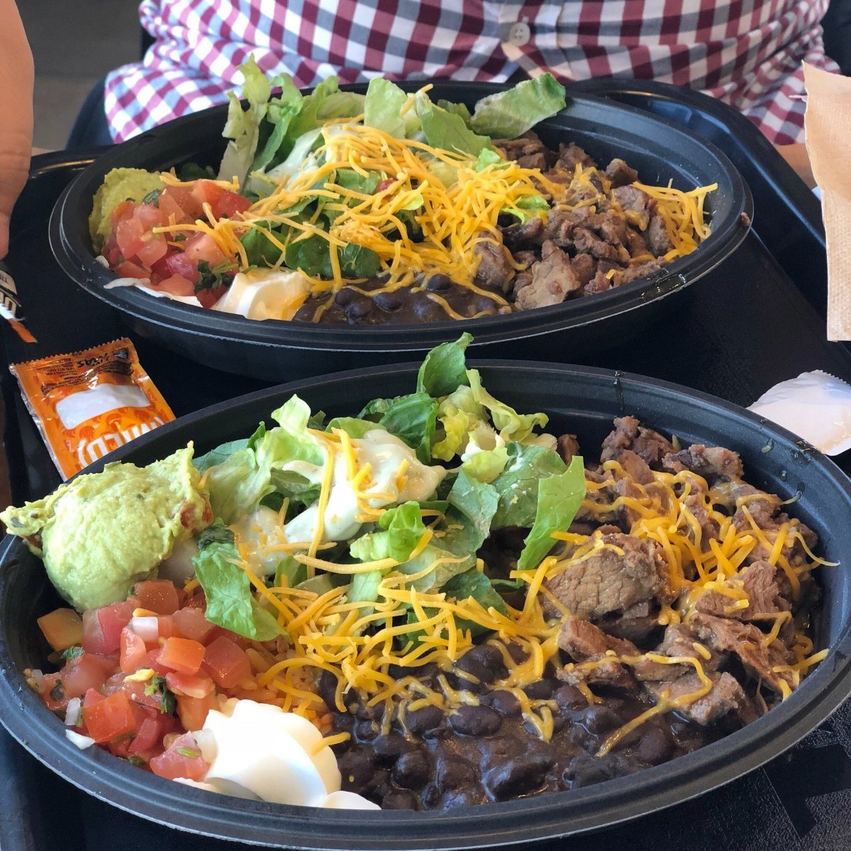 Two black bowls filled with burrito ingredients: shredded cheese, lettuce, diced tomatoes, guacamole, black beans, sour cream, and grilled beef. A packet of sauce and napkins are beside the bowls. A person in a plaid shirt is in the background.