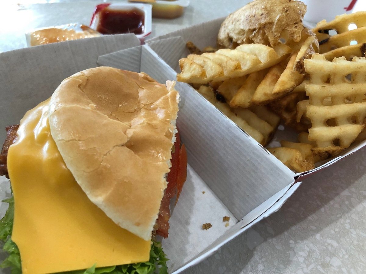 A sandwich with lettuce, tomato, bacon, and a slice of cheddar cheese next to a serving of waffle fries in a cardboard tray. Small containers of dipping sauce are visible in the background.