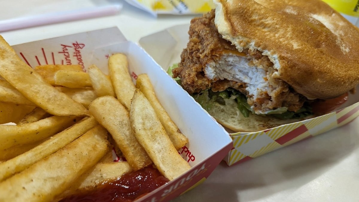 A close-up of a meal with crispy French fries next to a container of ketchup and a fried chicken sandwich on a bun with lettuce and tomato, served in a paper tray on a table.