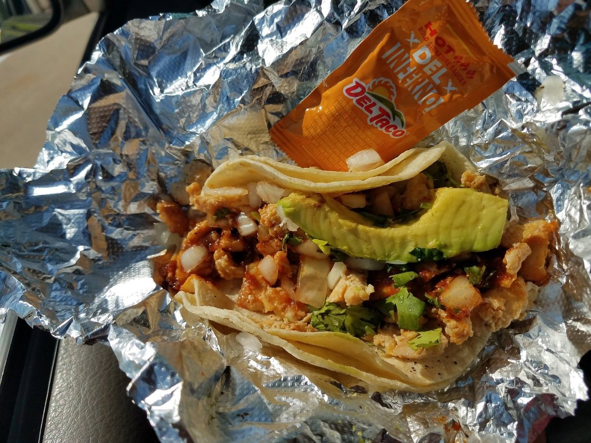 A taco with diced chicken, onions, cilantro, avocado, and hot sauce sits on foil. A Del Taco hot sauce packet is placed on top.