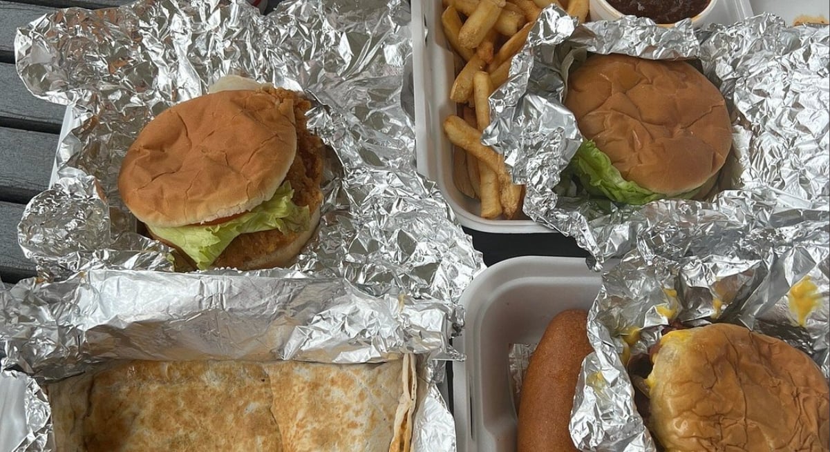 Four takeout meals in foil and white containers: two burgers with lettuce, fries with dipping sauce, a quesadilla, and a corndog. All items are arranged on a table, partially wrapped in foil or in open boxes.