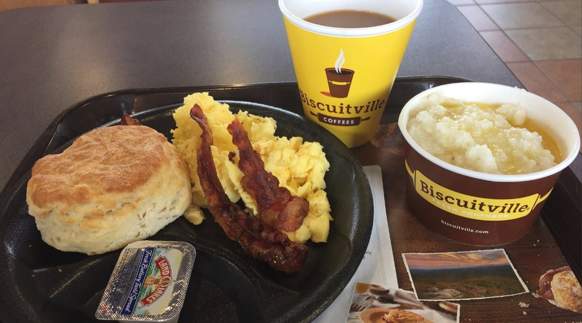 A breakfast tray with a biscuit, scrambled eggs, two strips of bacon, a cup of coffee, a bowl of grits, and a packet of grape jelly, all served in Biscuitville-branded containers.