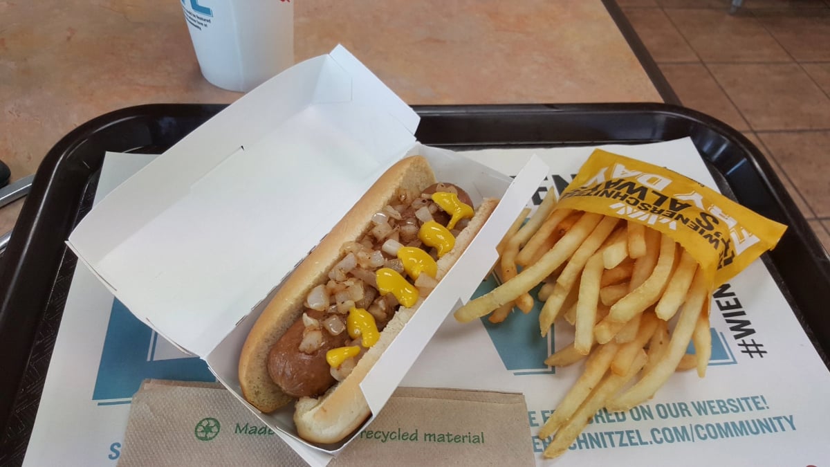 A hot dog topped with chopped onions and mustard in an open box next to a serving of French fries in a yellow paper pouch, all on a tray with a napkin and a drink cup in the background.