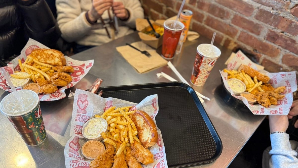 Three trays of fried chicken tenders, crinkle-cut fries, toast, coleslaw, and dipping sauce on a metal table, with drinks and people seated around the table at a casual restaurant.