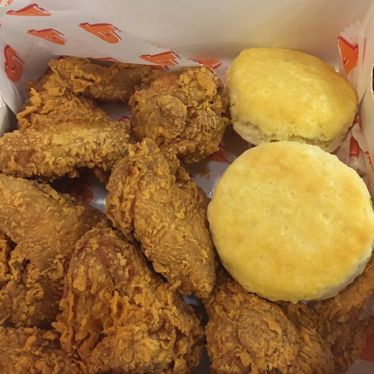 A box filled with pieces of crispy fried chicken and two golden brown biscuits on a sheet of printed paper.