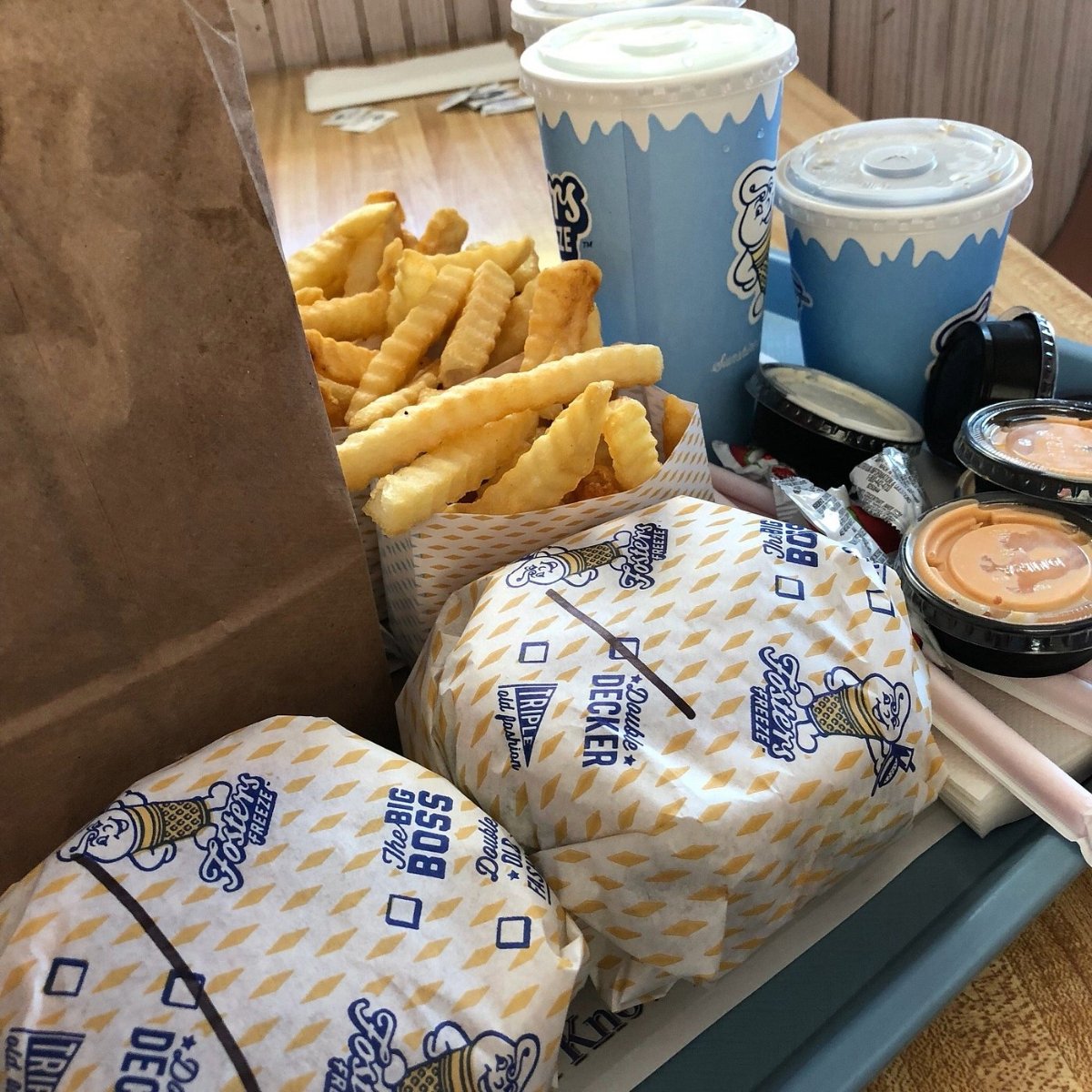 A tray with two wrapped burgers, a container of crinkle-cut fries, two blue soft drink cups with straws, sauce containers, and a brown paper bag on a wooden table at a fast food restaurant.