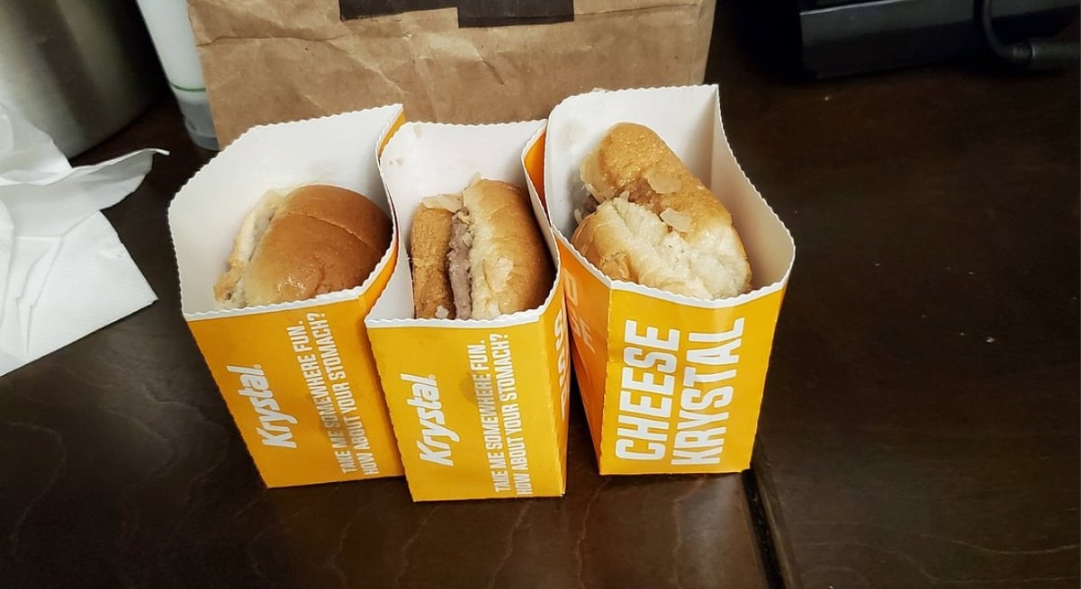 Three Krystal fast-food sliders in orange and white boxes are arranged side by side on a dark table. As one of the classic Southern fast-food chains, Krystal serves up classics like the Cheese Krystal, seen here with a brown paper bag in the background.