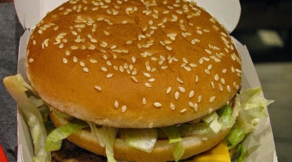 A close-up of a sesame seed hamburger bun with lettuce, pickles, a slice of cheese, and a beef patty, reminiscent of classic West Coast fast-food chains, served in an open cardboard container.
