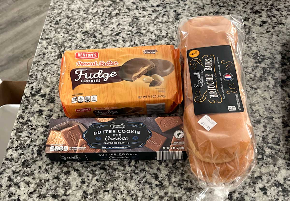Three packaged baked goods on a granite counter: Benton's Peanut Butter Fudge Cookies, Specially Selected Butter Cookie with Chocolate, and a loaf of Specially Selected Brioche Buns.