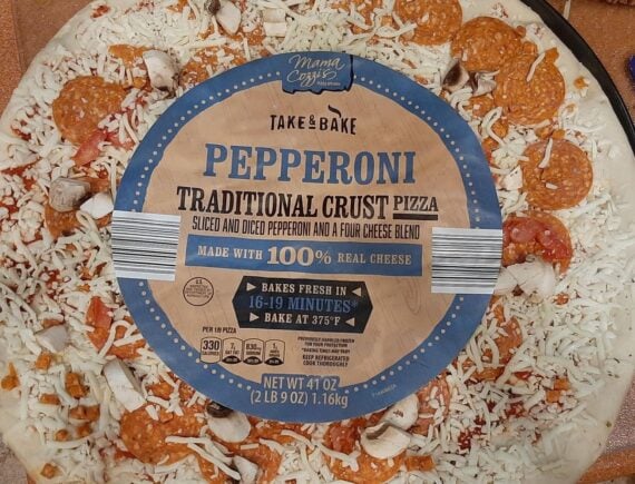 A take-and-bake Mama Cozzi&rsquo;s pepperoni traditional crust pizza with sliced and diced pepperoni, mushroom pieces, and shredded cheese, shown in its packaging with a blue label.