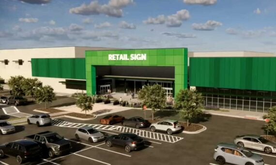 A modern Amazon store with a green facade labeled "Retail Sign," large windows, and an inviting entrance area. Several cars fill the parking lot, while people walk toward the entrance. Trees line the parking area for added greenery.
