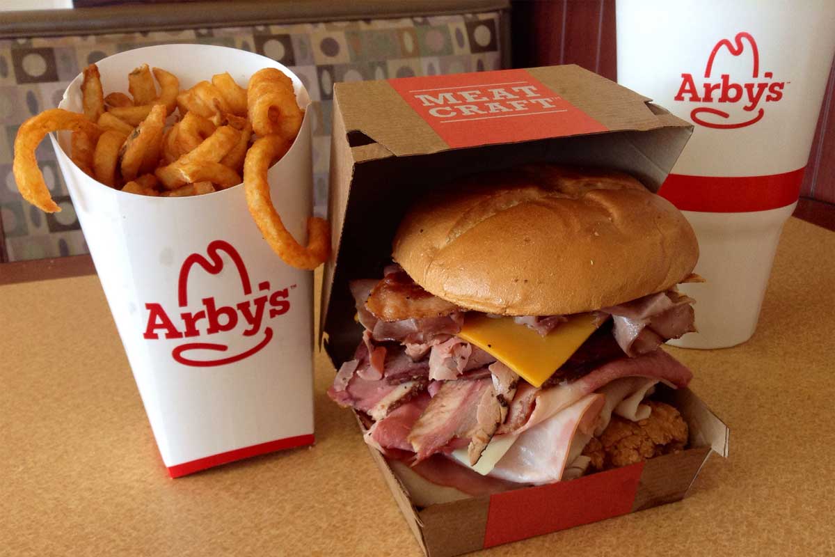 A meal from Arby’s featuring a large cup of curly fries, a stacked roast beef sandwich with cheese in a cardboard box, and a large Arby’s soft drink, set on a table in a restaurant booth.