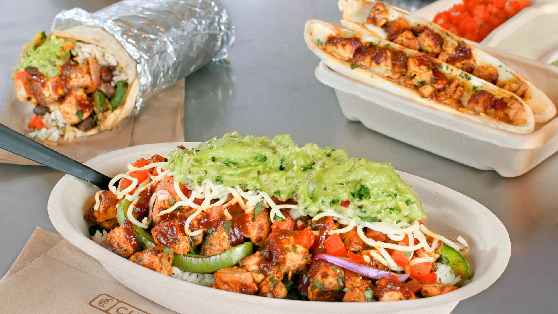 A burrito bowl with chicken, rice, vegetables, cheese, pico de gallo, and guacamole sits in the foreground—part of a new fast-food menu. In the background, a wrapped burrito and two soft tacos with chicken and toppings are on a table.