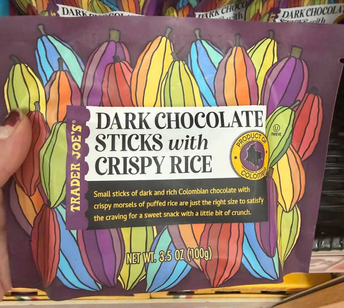 A hand holds a bag of Trader Joe's Dark Chocolate Sticks with Crispy Rice. The purple package features colorful illustrations of cocoa pods and product details in white and yellow text.