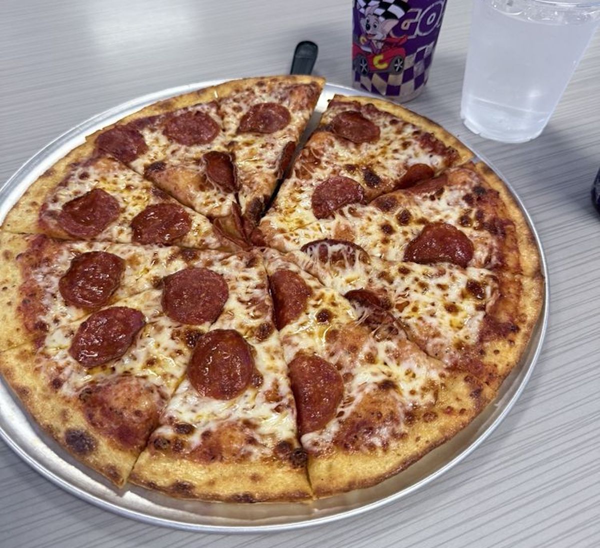 A pepperoni pizza cut into eight slices sits on a metal tray next to a cup with a purple cartoon design and a clear plastic cup of water on a light-colored table.