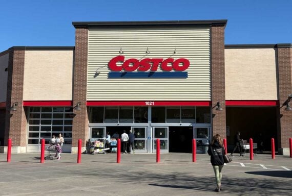 People walk toward the entrance of a Costco warehouse store on a sunny day. The building features the large red Costco logo above the doors, with several shopping carts and red bollards out front.