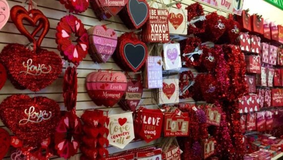 A store display filled with various Valentine&rsquo;s Day decorations, including red and pink heart-shaped wreaths, signs with messages like &ldquo;Love&rdquo; and &ldquo;Happy Valentine&rsquo;s Day,&rdquo; and glittery ornaments hanging on hooks.