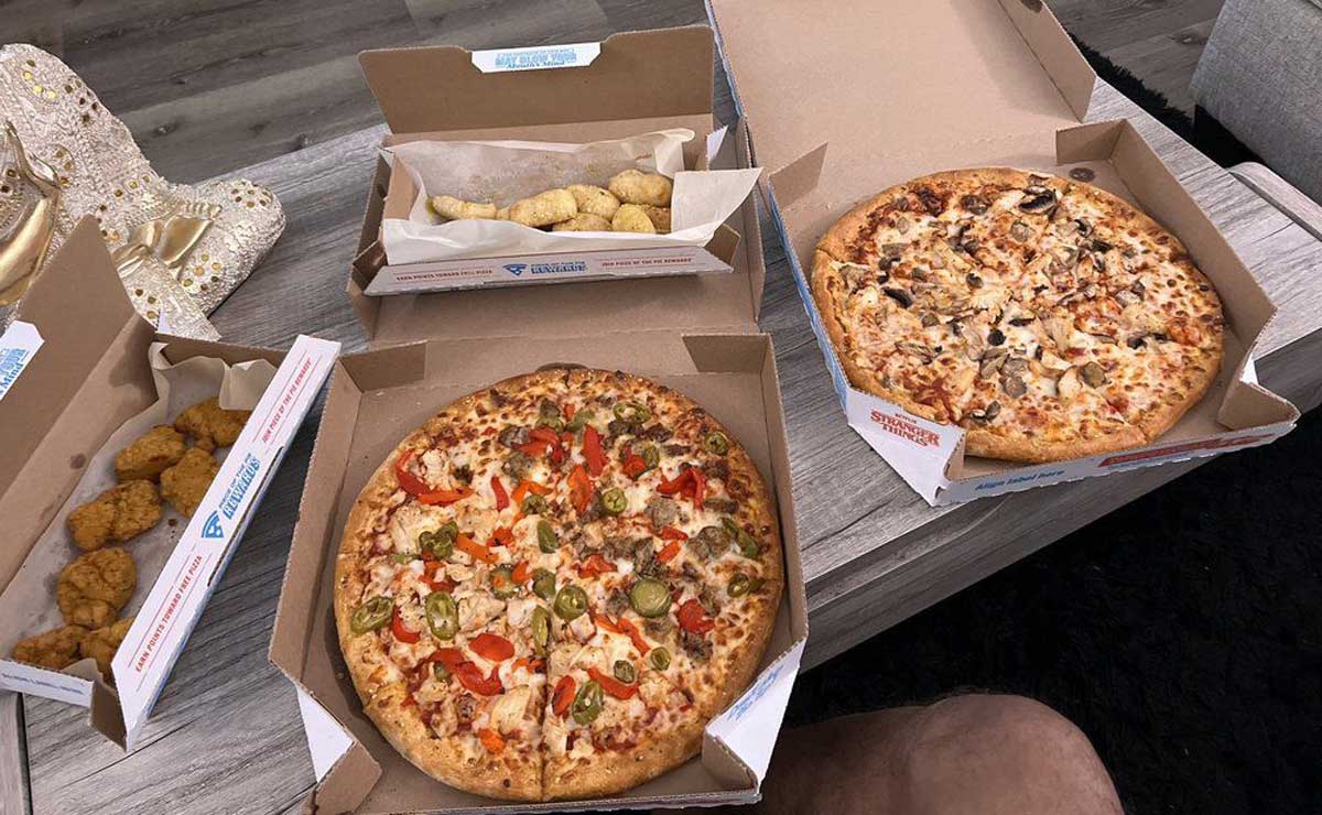 Two open pizza boxes with different pizzas, one topped with vegetables and meat, the other with mushrooms and chicken. Sides of chicken bites and bread twists are also in open boxes on a wooden table.