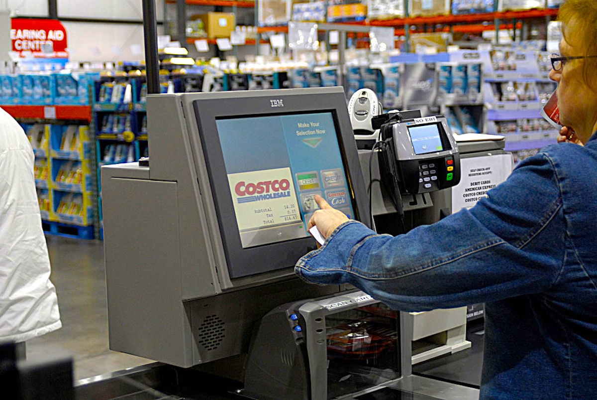 A person uses a touchscreen self-checkout kiosk at Costco, with a credit card terminal and store shelves visible in the background.