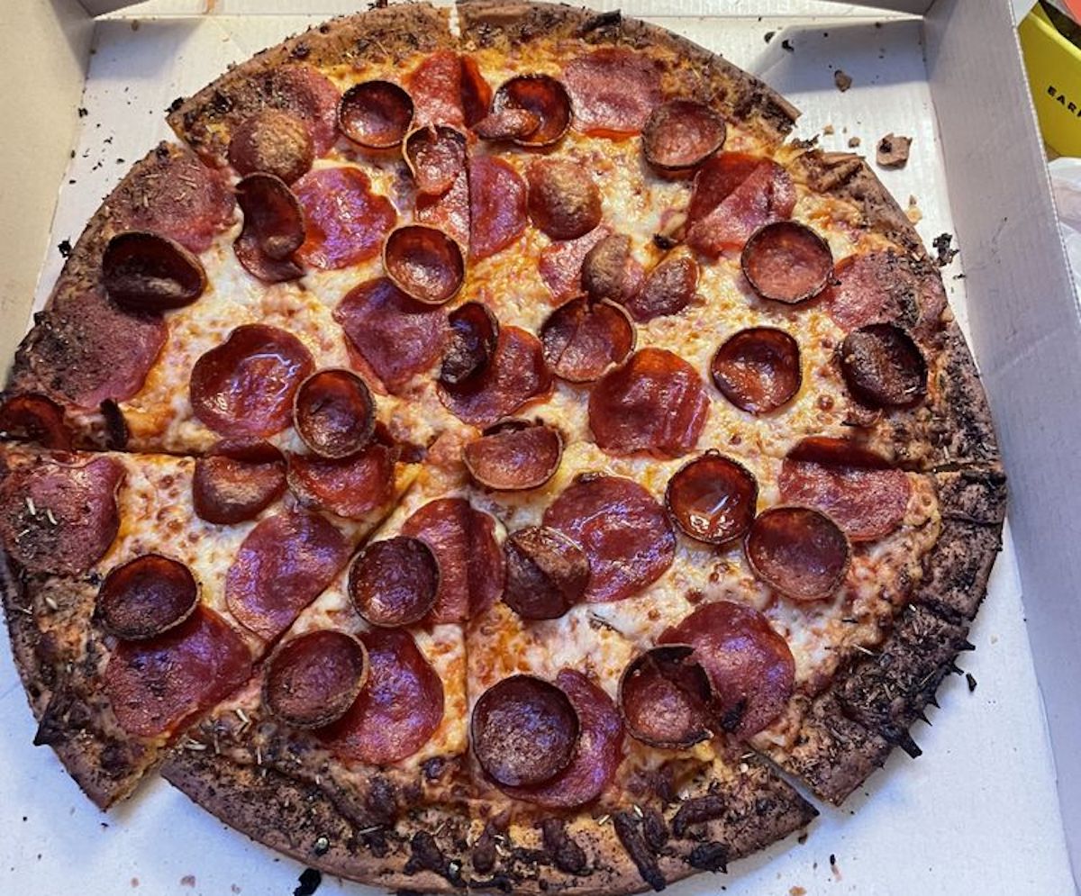A large pepperoni pizza with a well-done, slightly burnt crust sits inside an open pizza box. The pizza is sliced into eight pieces and is topped with melted cheese and pepperoni slices.
