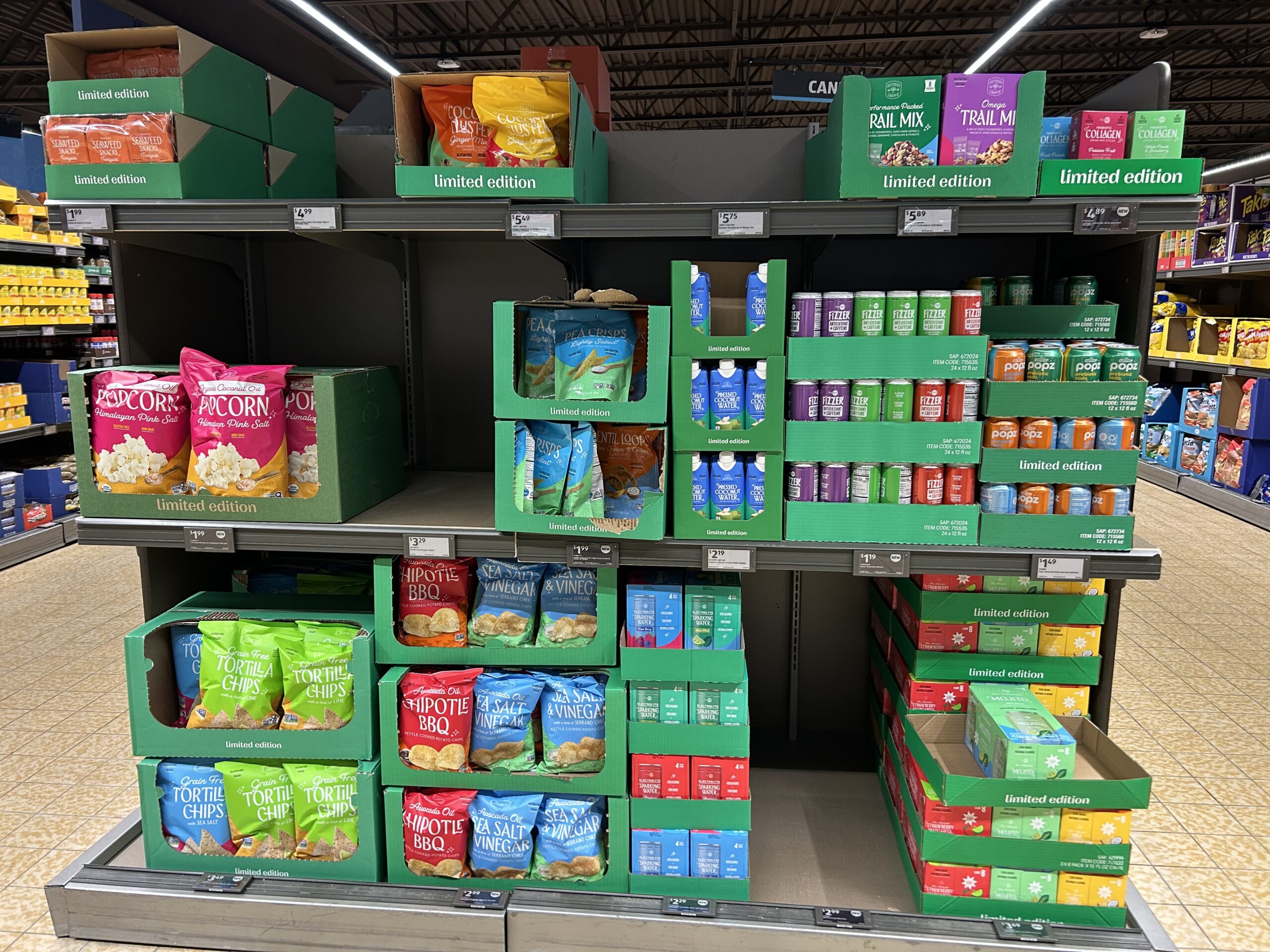 A supermarket shelf displays limited edition snacks, including popcorn, trail mix, and various flavored tortilla chips, neatly arranged in green boxes with some sections partially empty.