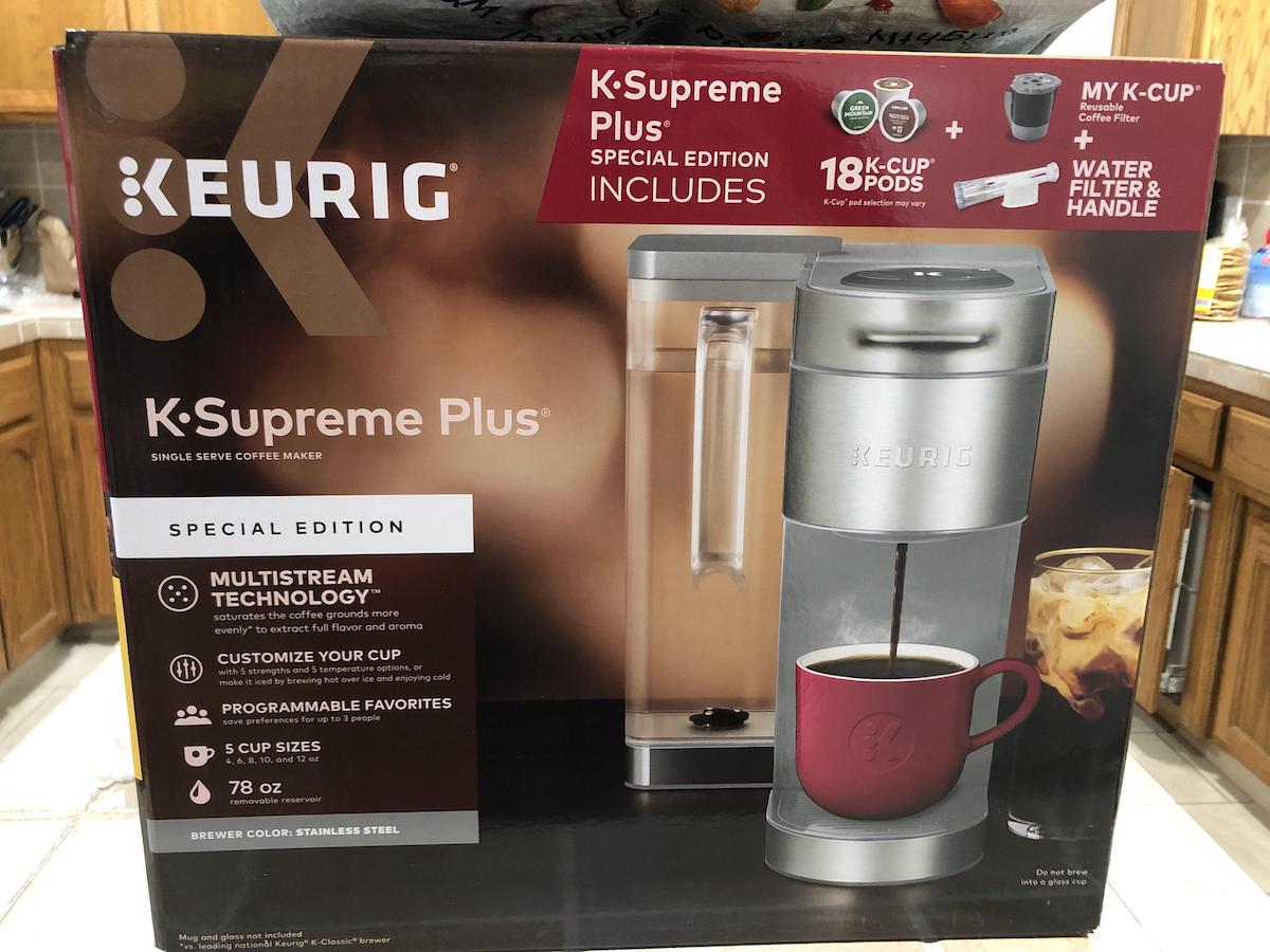 A Keurig K-Supreme Plus coffee maker in its box, featuring the machine, a red coffee mug with coffee, and promotional text highlighting included K-Cup pods, a water filter, and the My K-Cup accessory.