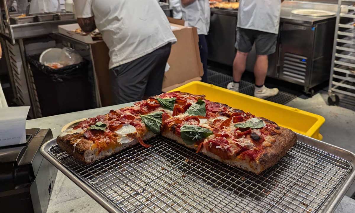 A rectangular pepperoni pizza, reminiscent of classic East Coast cheap pizza slices, sits on a cooling rack in a commercial kitchen, with chefs working in the background and stainless steel equipment visible.