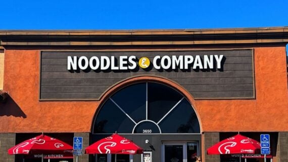 Storefront of Noodles & Company with its sign above the entrance, two red patio umbrellas, two accessible parking signs, and a bright blue sky in the background.