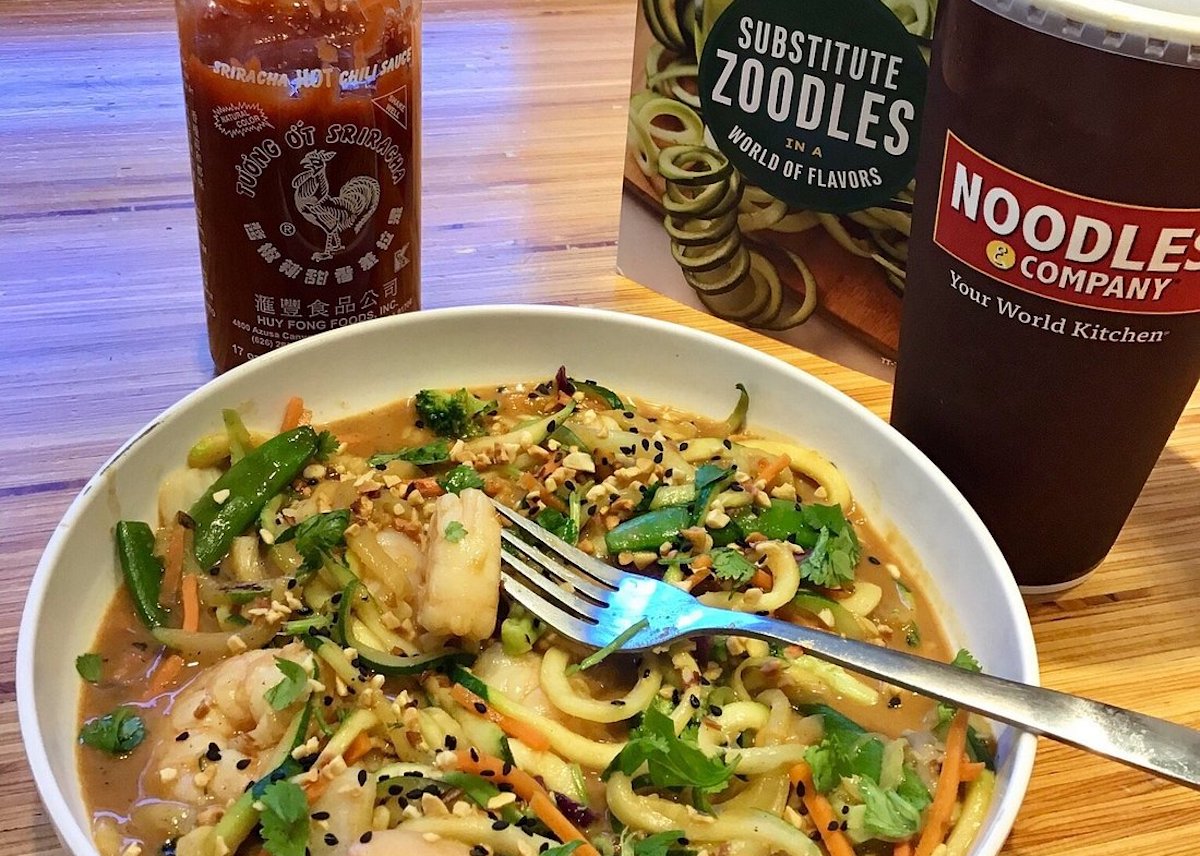 A bowl of noodle stir-fry with shrimp, vegetables, sesame seeds, and herbs, next to a Sriracha bottle, a Noodles & Company drink cup, and a sign promoting zoodle substitutions. A fork rests in the bowl.