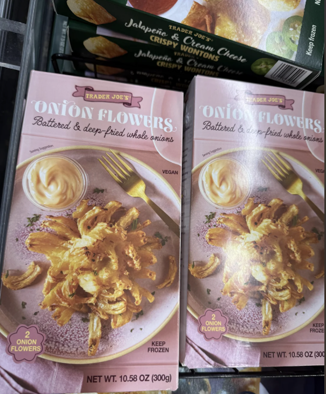 Two boxes of Trader Joe’s Onion Flowers, battered and deep-fried whole onions, are displayed in a freezer. The packaging shows a plated onion flower with dipping sauce and a fork on a light pink plate.