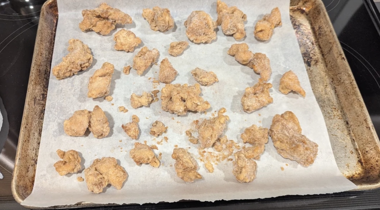 Small, breaded pieces of chicken are spread out on a parchment-lined baking sheet, ready to be cooked or freshly baked. The baking sheet has some browned edges, and the chicken pieces appear crispy.