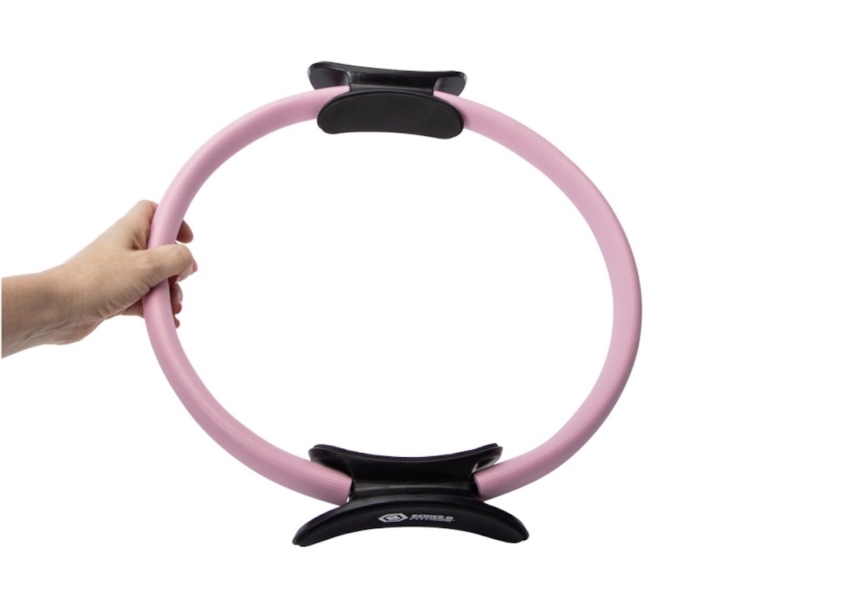 A hand holding a pink Pilates ring with black padded grips on the top and bottom, against a plain white background.