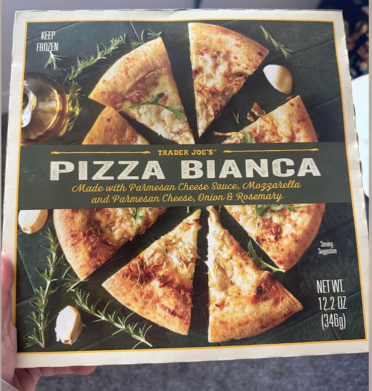 A hand holds a box of Trader Joe’s Pizza Bianca, featuring a photo of a sliced pizza topped with cheese, onion, and rosemary. The box highlights mozzarella, parmesan, and a net weight of 12.2 oz (346g).