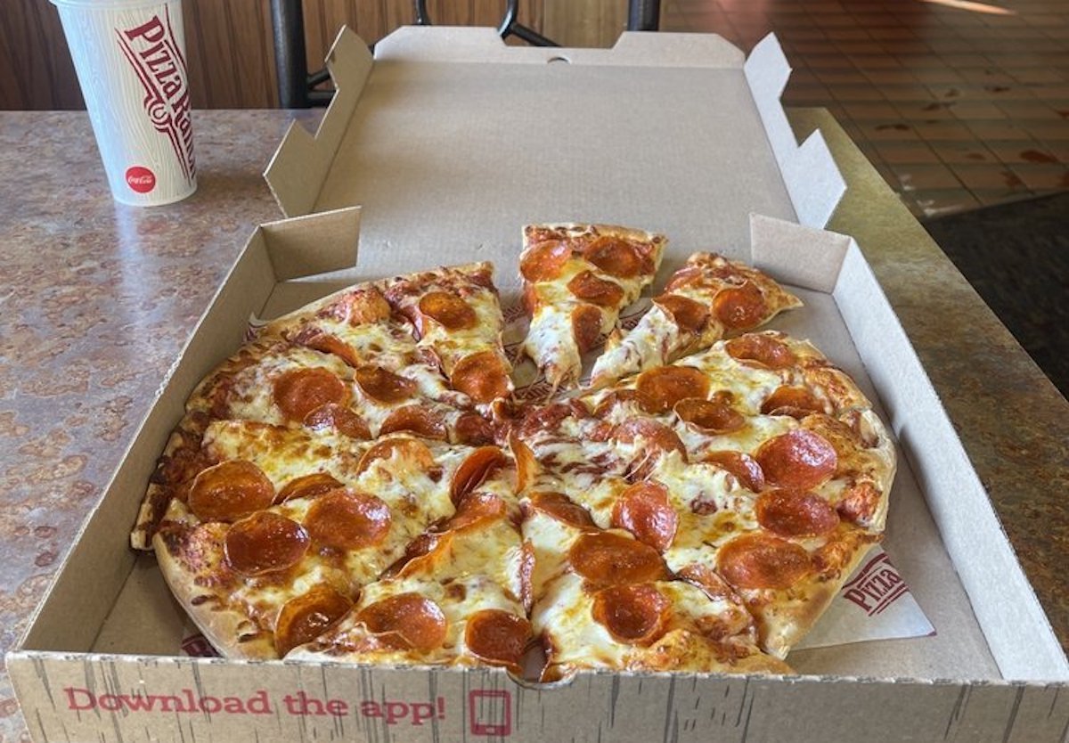 An open pizza box on a table contains a pepperoni pizza with melted cheese and nine slices. A disposable cup with a straw sits in the background. The table and floor have a brown, tiled pattern.