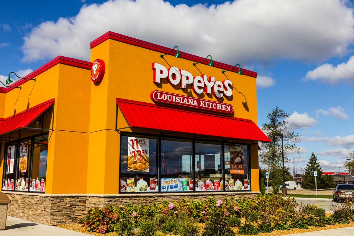 A Popeyes Louisiana Kitchen restaurant with bright orange and yellow exterior, red accents, and large logo, set under a blue sky with scattered clouds. Menu posters are visible in the windows.